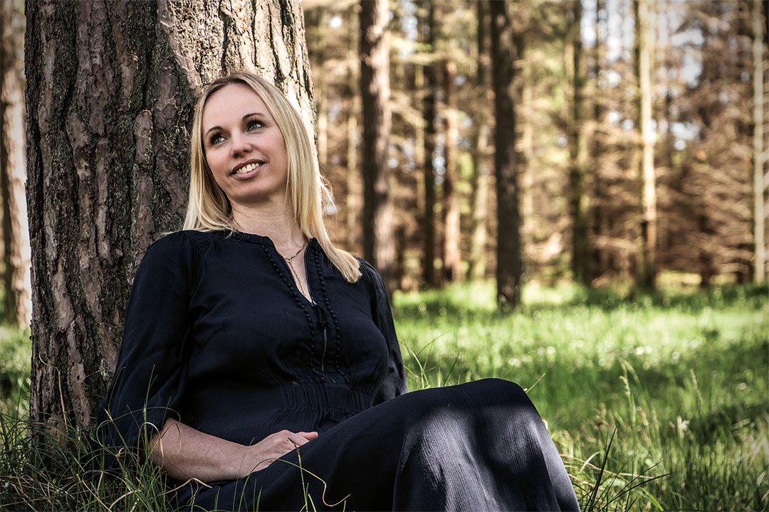 Portrætfoto af Fysioterapeut Psykoterapeut og TRE behandler Louise Møldrup der sidder i en blå kjole op ad et træ i en skov
